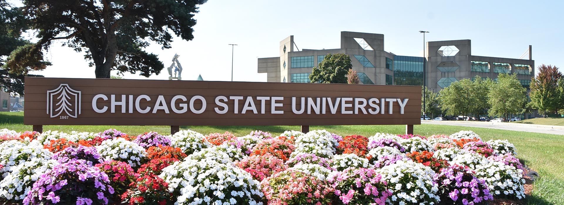 Chicago State University entrance sign surrounded by colorful flower beds, with Gwendoly Brooks Library and trees in the background.