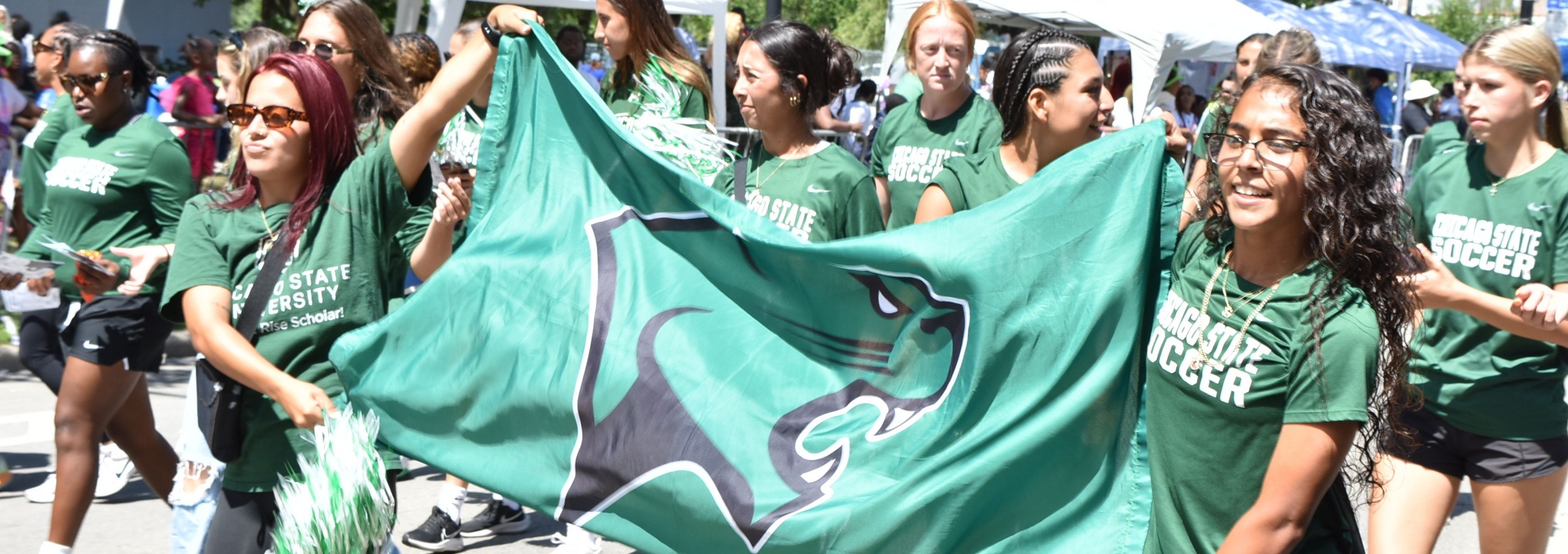 Chicago State University women's soccer team walking in a parade while holding a large, green CSU mascot flag.