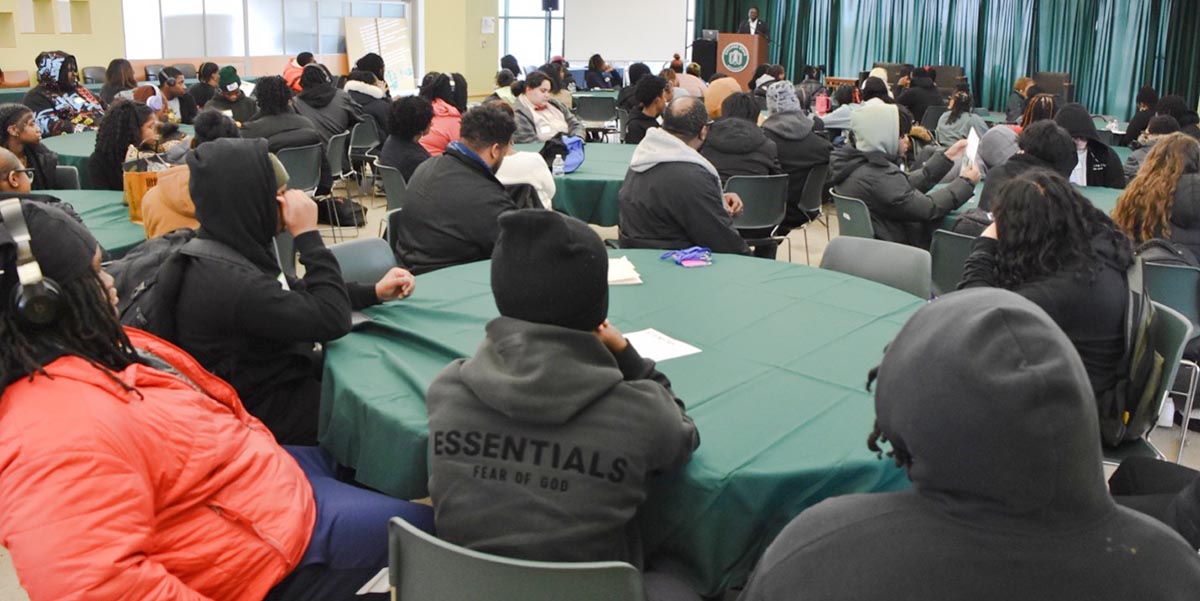 A large group of people seated at round tables as someone speaks at a podium during STEM-Day Hackathon