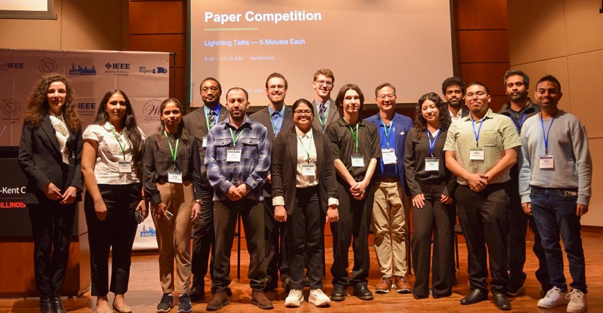 A group of participants wearing lanyards with name badges stand on stage in a warmly lit conference room.