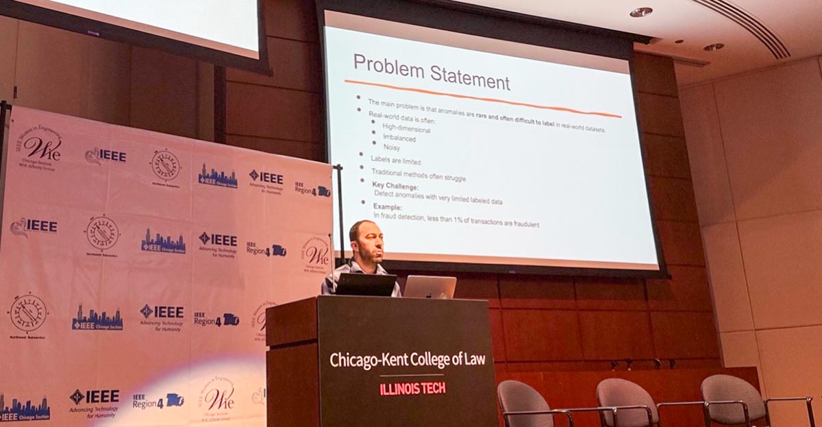 Ozgur Gurbuz, Chicago State University CIMST student, stands at a podium at the Chicago-Kent College of Law, giving a presentation in front of a large screen. The slide shown is titled "Problem Statement," and the background wall behind the speaker is covered in a banner featuring a step-and-repeat of IEEE (Institute of Electrical and Electronics Engineers) and 'Women in Engineering' (WIE) logos.