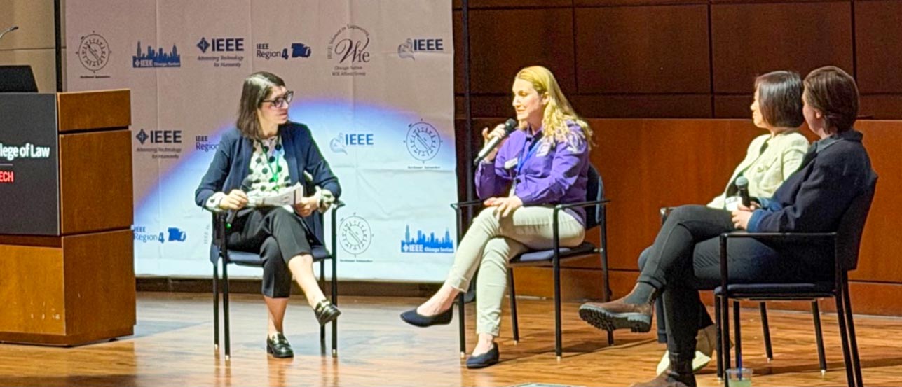 A group of four panelists sit on a brightly lit stage, with one woman in a purple shirt speaking into a microphone while the others listen.
