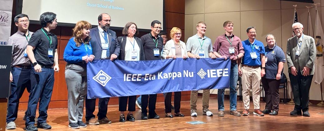 A group of conference participants stand on stage holding a blue fabric banner that reads "IEEE-Eta Kappy Nu."