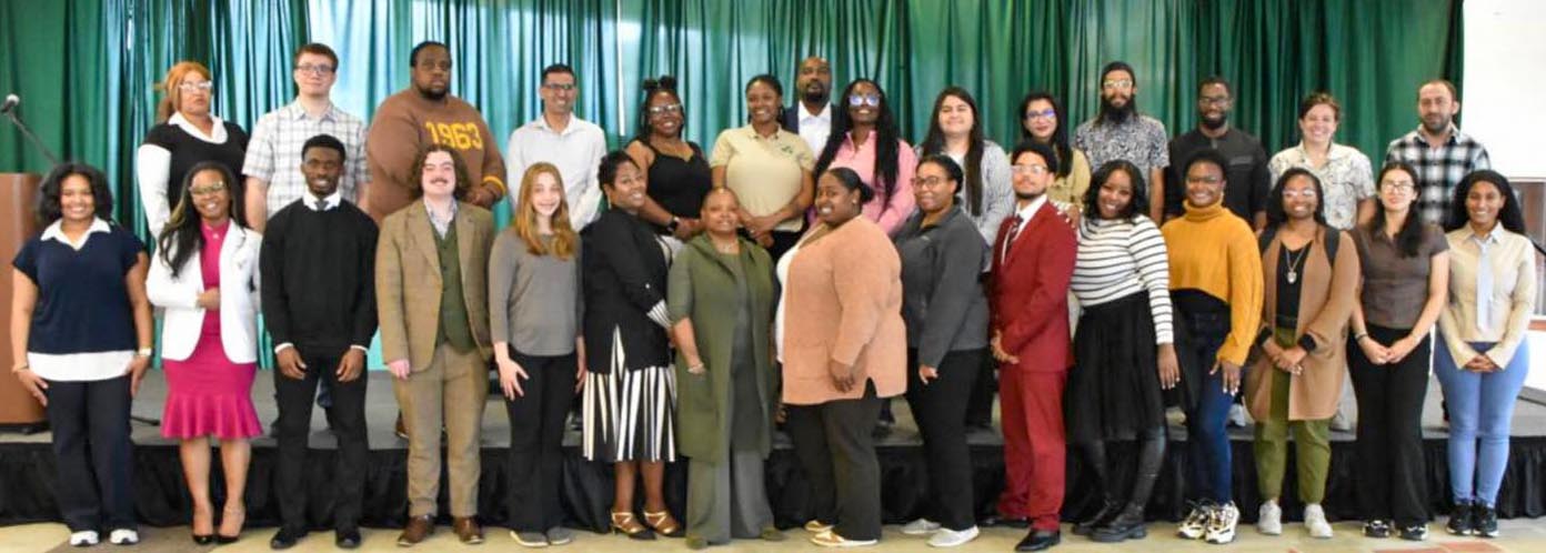 A group of approximately 30 student researchers pose together on stage in front of a green backdrop.