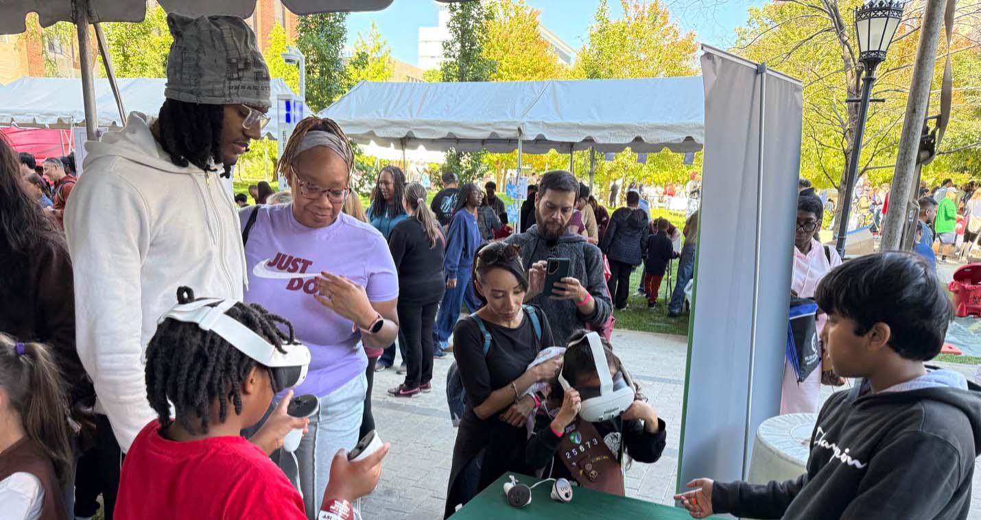 A group of people stand next to Chicago State's table outside while two young children try out virtual reality headsets. A crowd of festival attendees can be seen in the background.