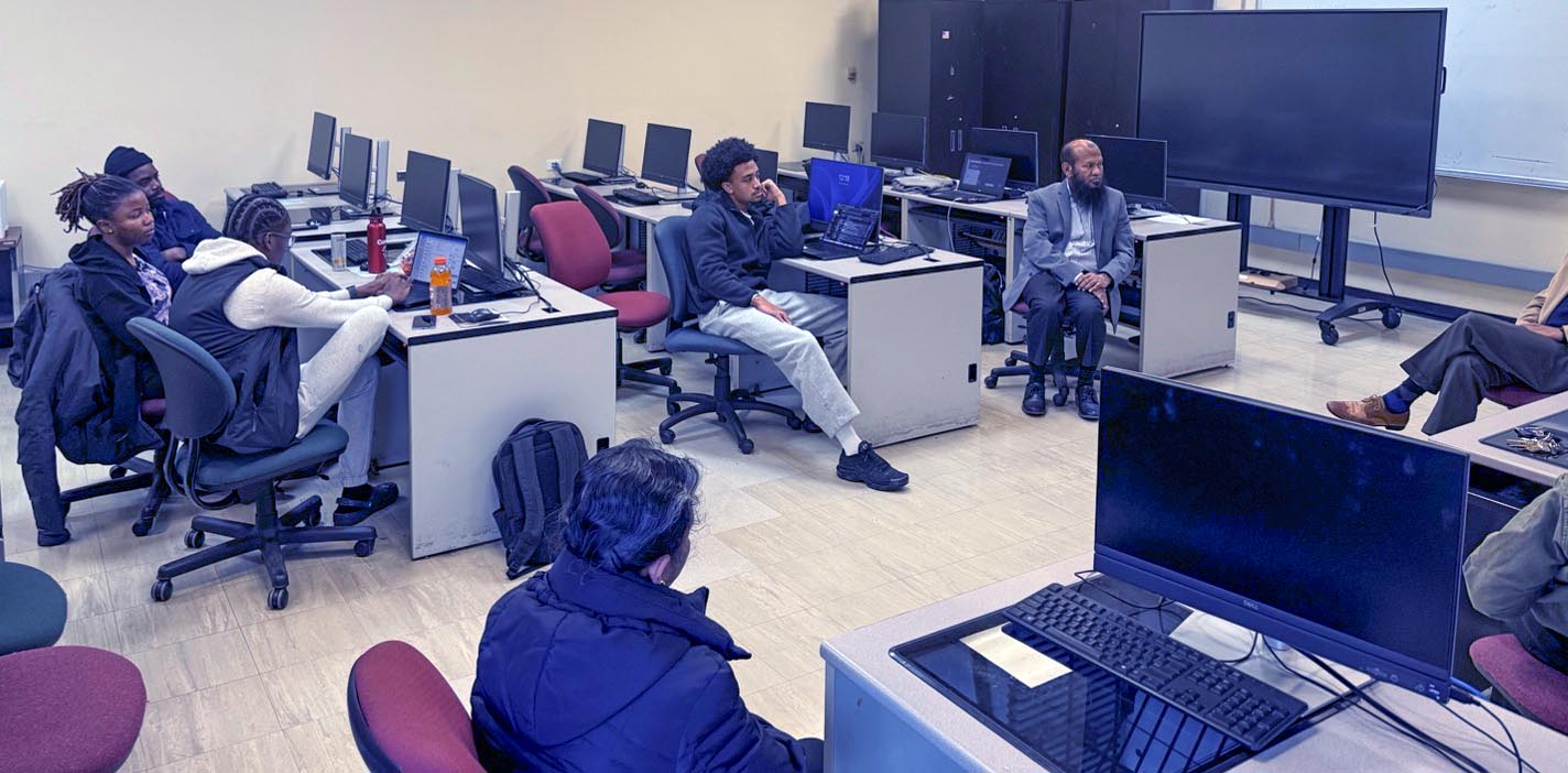 A group of students and a faculty member sit in a computer lab, listening while someone speaks.