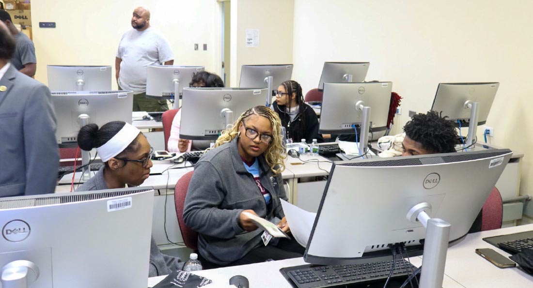 Three high school students sit next to each other in a computer lab with large monitors.