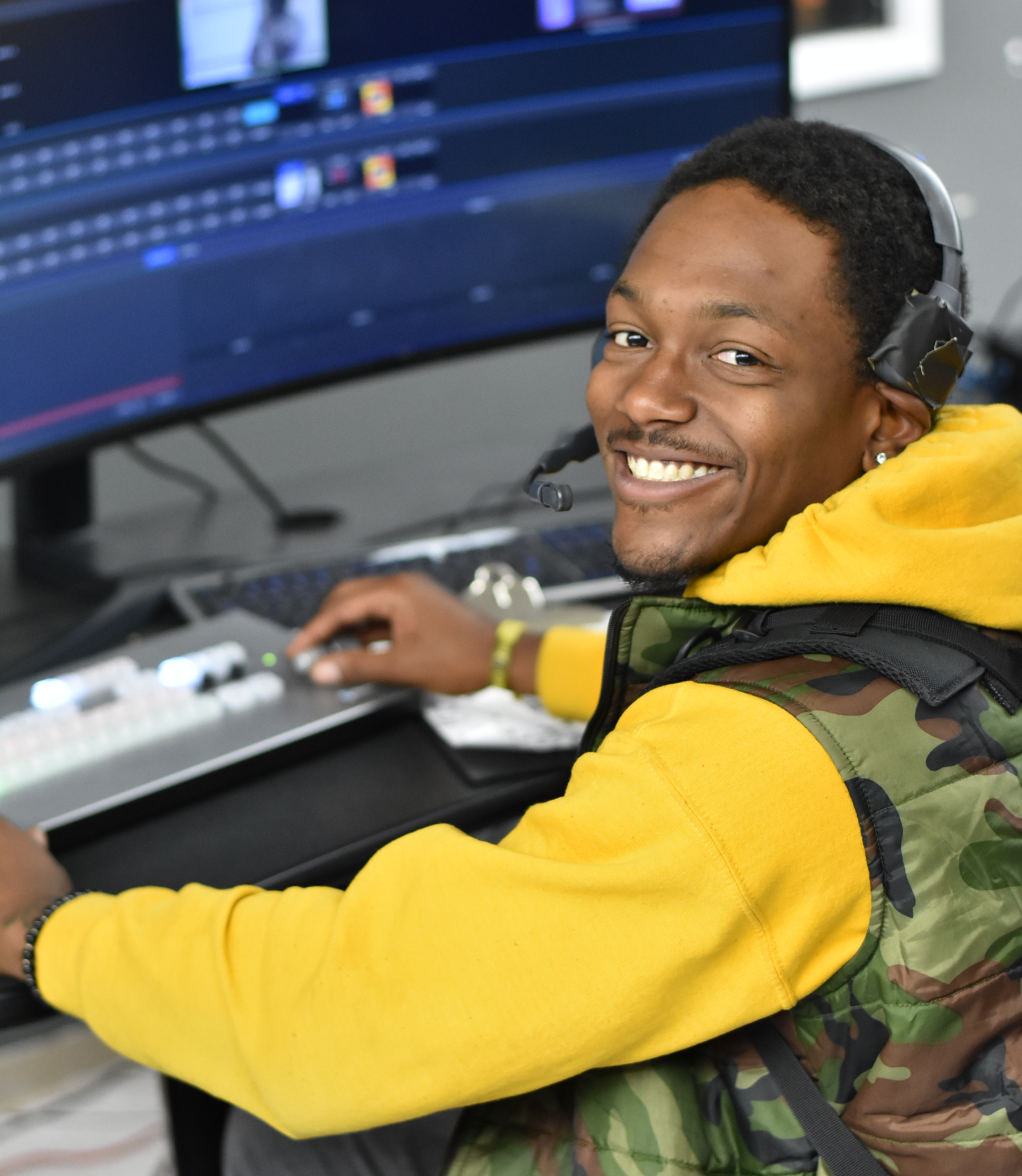 A young man wearing a headset and yellow hoodie smiles at the camera while working at a computer workstation with video editing software.