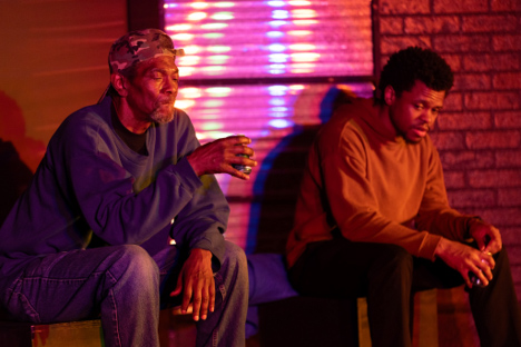 Two men seated next to each other on a dimly lit stage illuminated with warm red and purple theatrical lighting during a CSU theatre performance.