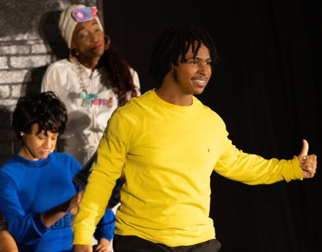 A CSU theatre stage performance scene featuring a young man in a yellow long-sleeve shirt in the foreground, with two other performers behind him in colorful outfits.