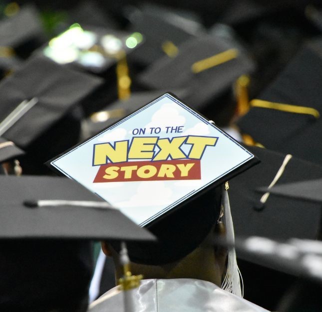 A close-up view of a graduate's mortarboard featuring a "Toy Story" inspired design that reads "On to the Next Story" against a blue sky and cloud background.