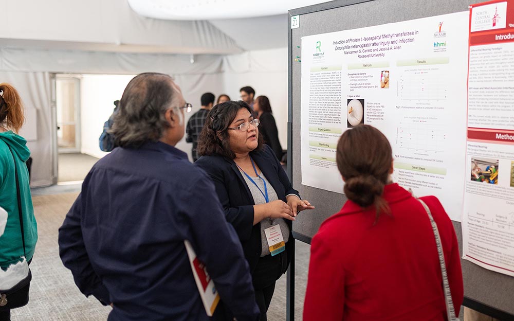 A student with short, brown hair and a black blazer with a gray blouse presents her research poster to two listeners. In front of them is a large poster.