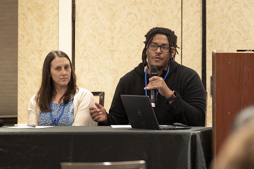 Two panelists, a man and a woman, seated side-by-side at a table covered with a shiny, black tablecloth. The man, with dreadlocks and glasses, is speaking into a microphone. An open black laptop sits on the table in front of him. The woman to his left is listening attentively.