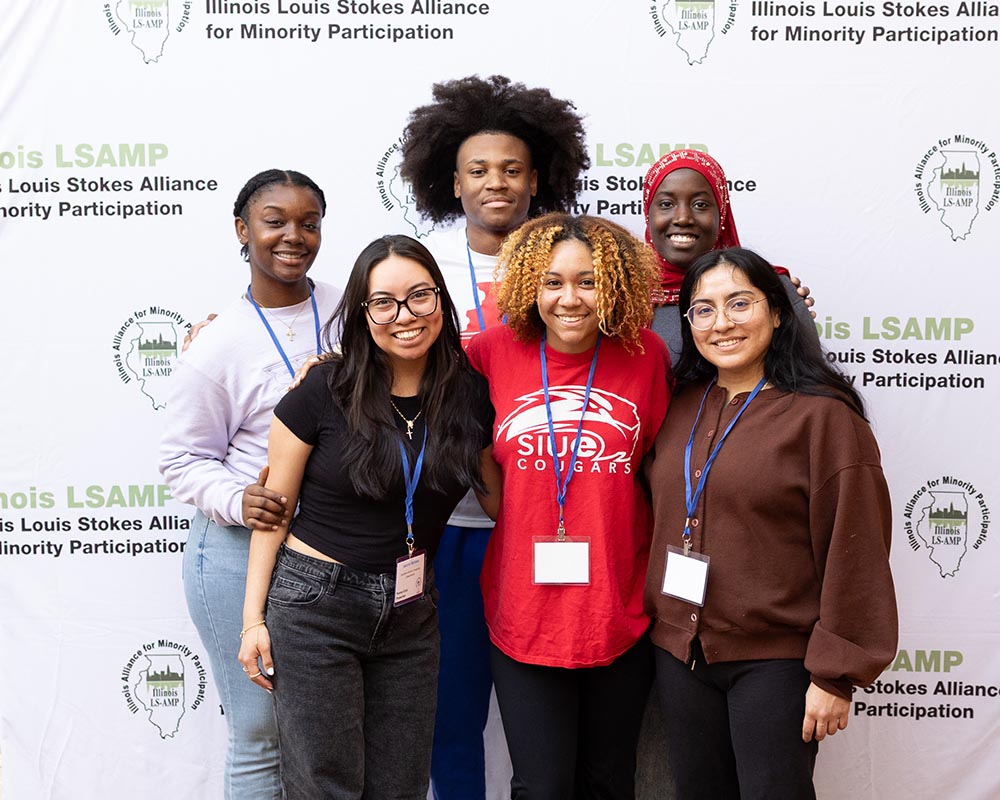 Six students smile and stand closely together to pose for a photo in front of a white backdrop with the Illinois LSAMP logo across it.