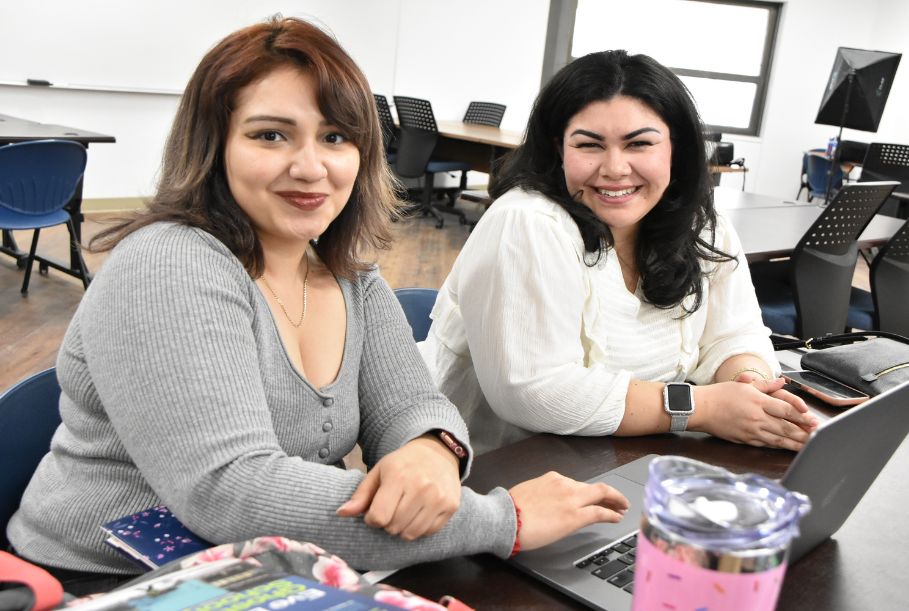 Two women sitting at a table in a computer lab look up from their laptops and smile at the camera.