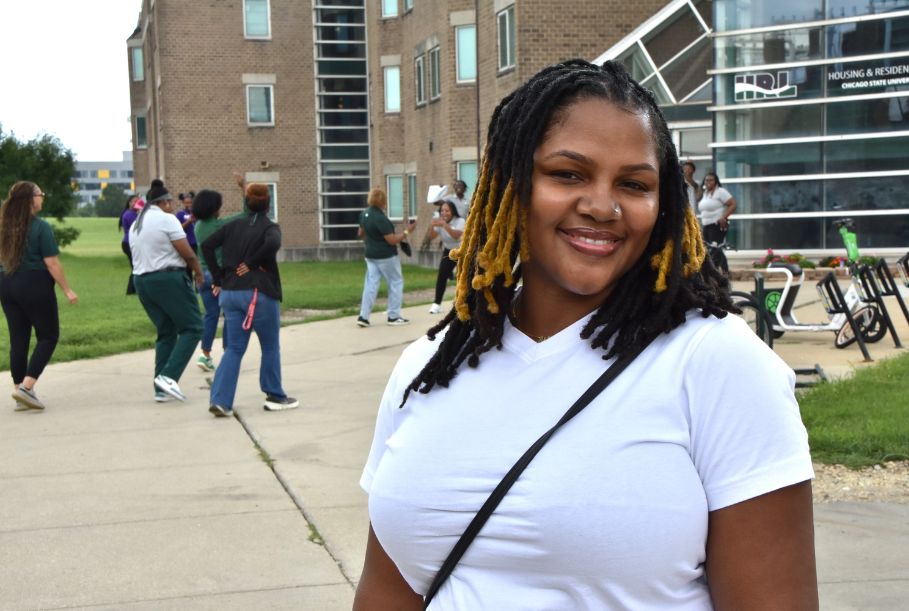 A woman smiles into the camera, standing in front of Chicago State University’s residence hall while students walk and socialize in the background. She has dark brown and blonde dreadlocks and is wearing a white t-shirt with a black cross-body bag. 