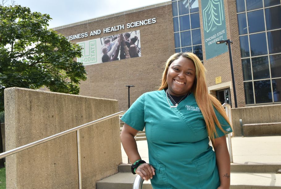 A student in teal Chicago State University scrubs poses on the stairs outside the Business and Health Sciences building, which features large university banners and glass windows.