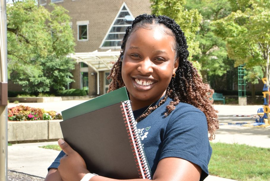 A sunny photo of a woman in a dark, blue CSU shirt smiling into the camera, hugging her notebook and folder outside of the Cordell Reed Student Union Building.