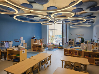 A pre-k classroom with a calming blue color scheme and modern circular lighting. Large windows let in natural light and low shelving units are filled with learning materials, books and toys.