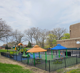 photo of the playground from another angle, featuring slides, covered picnic tables with orange and blue roofs and the College of Education building and trees in the background
