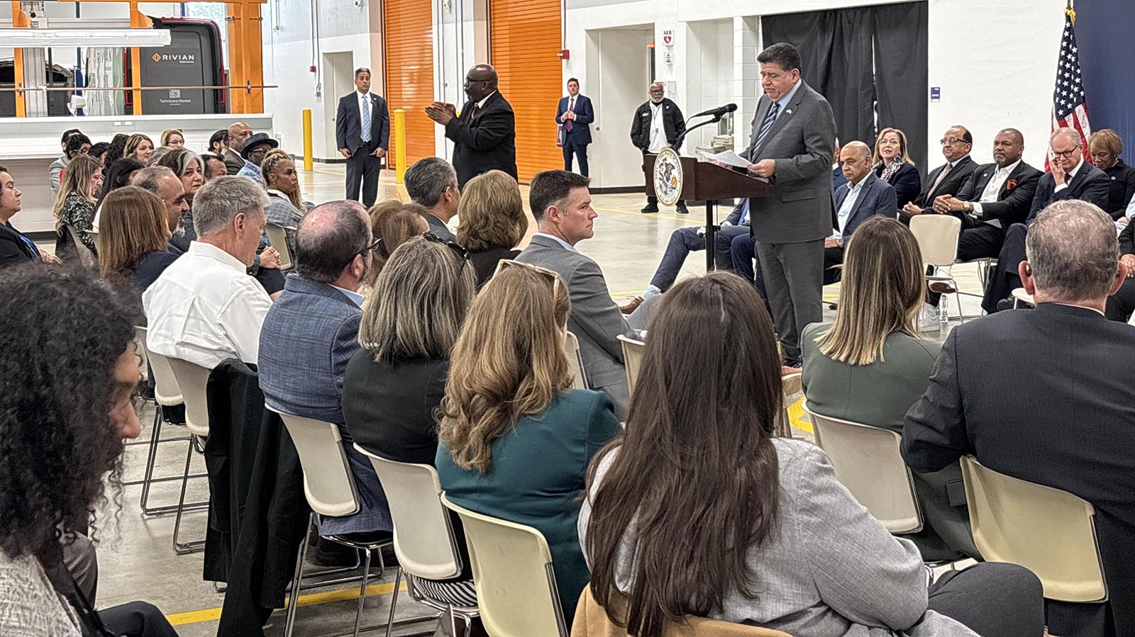 Illinois Governor JB Pritzker stands at a podium and addresses a seated audience in a brightly lit industrial garage