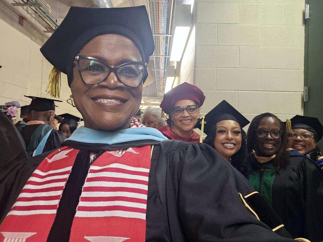 A woman wearing a doctoral robe and cap with a red stole and glasses smiles for a selfie with Dr. Man and graduates behind her in a large hallway with while other graduates get ready for commencement.
