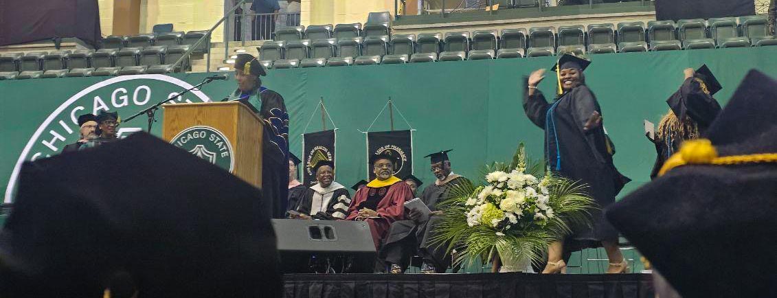 A proud social work graduate smiles and waves as she walks across the stage to receive her diploma. There is a large white flower arrangement at the edge of the stage and administrative leaders sitting toward the back of the stage. Behind them is a long green backdrop with the presidential seal of Chicago State University.