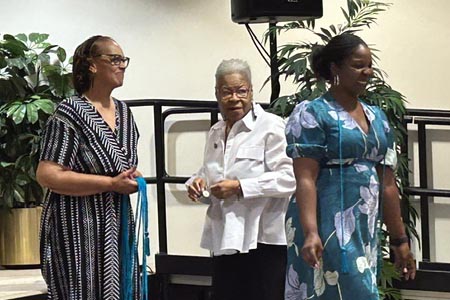 Three women standing on a stage during a ribbon pinning ceremony. On the left, the social work department chair, Dr. Mann, wears glasses and a patterned black and white dress, smiling while holding a bundle of blue cords. In the center, an older woman with short gray hair and a white button-down shirt looks toward the right. On the right, a woman wearing a teal floral dress walks across the stage with a blue cord draped over her shoulder.