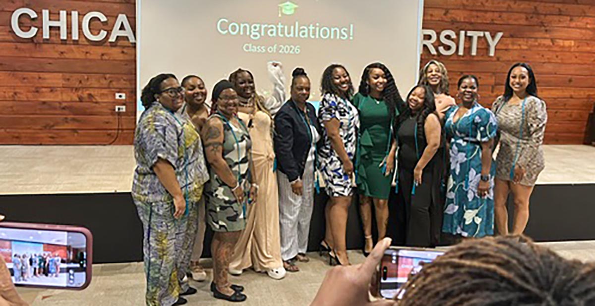 A group of eleven smiling women stand together on a stage for a graduation group photo with most wearing teal graduation honor cords. They are positioned in front of a large projection screen displaying a green graduation cap icon and the text "Congratulations! Class of 2026."