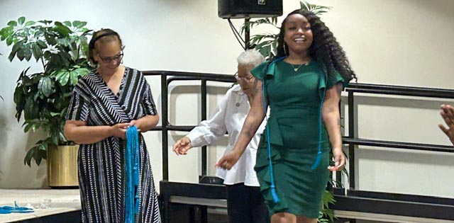 Three women standing in front of a stage. A woman with long hair wearing a green dress walks away with a smile after receiving her teal blue honor cord. Dr. Mann, chair of the social work deparment, looks down at the bundle of blue cords in her hands while an older woman with gray hair stands next to her, offering a hand of assistance.